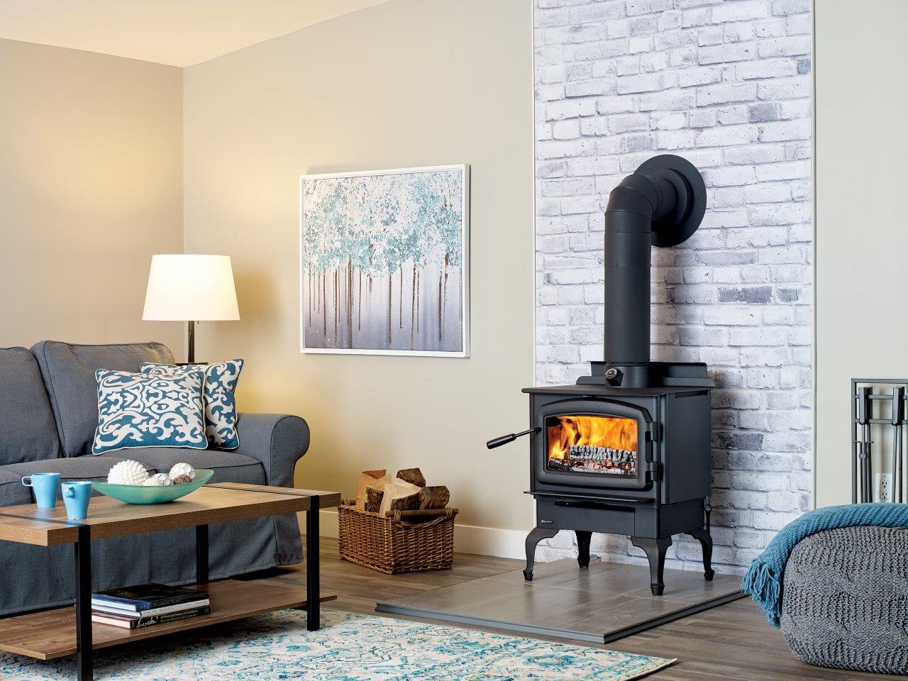 Cozy living room with a lit wood-burning stove against a white brick wall. A gray sofa, coffee table, and art complete the scene.
