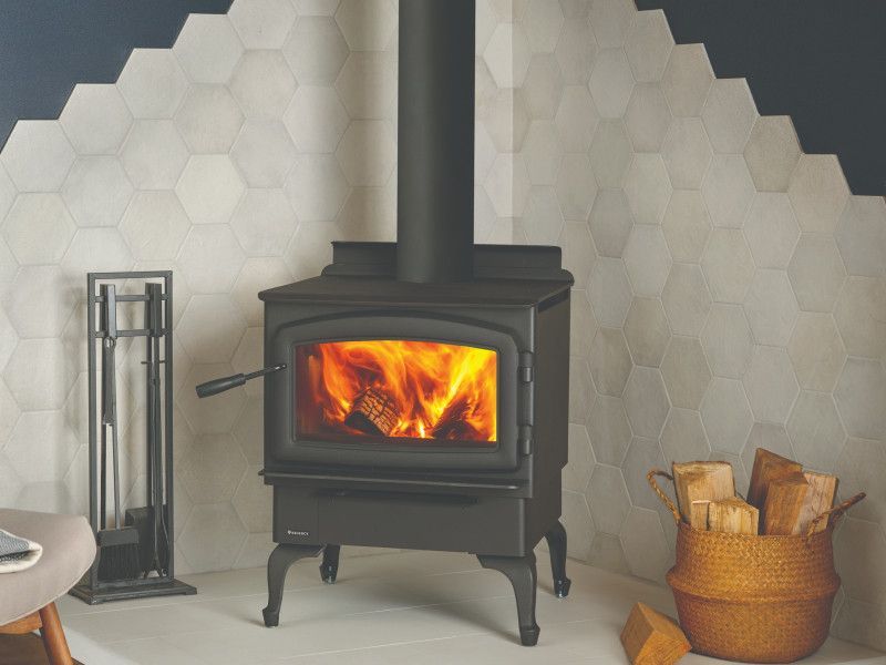A black wood-burning stove with a roaring fire, set against a wall with white honeycomb tiles. A basket of wood and a fireplace tool set are nearby.