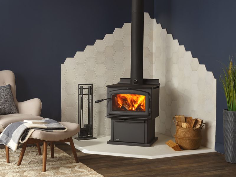 A black wood-burning stove in a corner setting, with a lit fire. Adjacent to it are firewood, a decorative tool set, and a comfortable seating area.