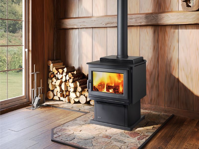 A burning wood stove in a rustic wooden cabin, with a stack of firewood and a window overlooking a landscape.