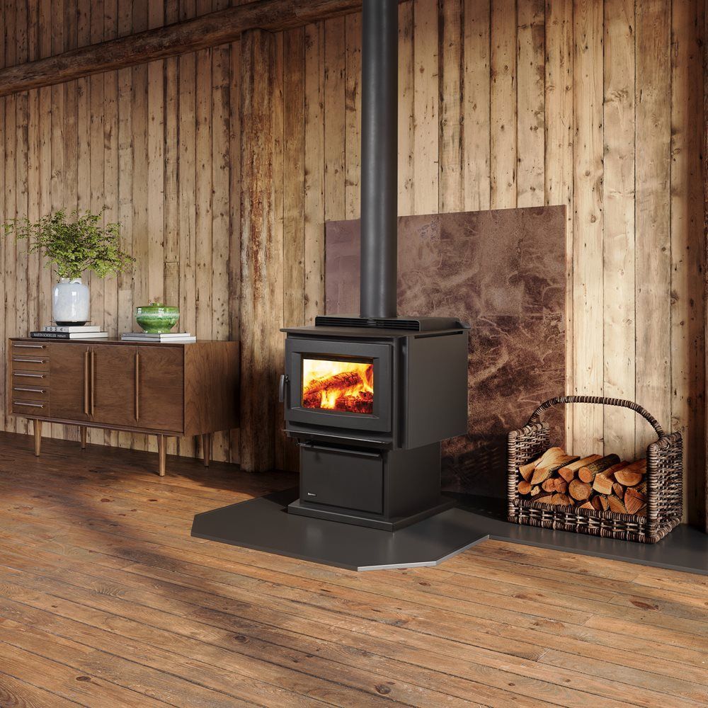 A black wood-burning stove with fire in a room with wooden walls and floor, next to a basket of firewood and a retro cabinet.
