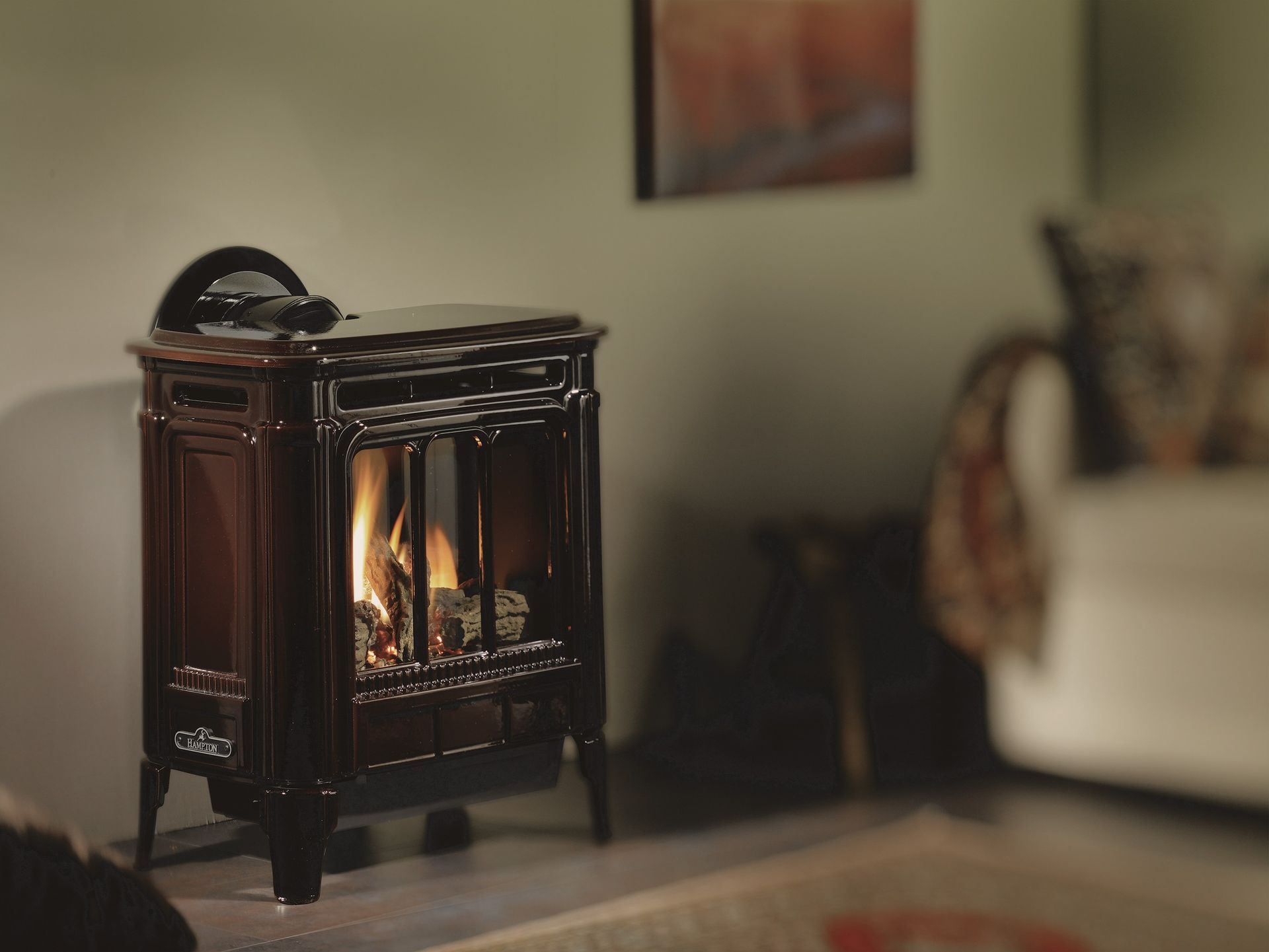 A dark brown gas stove with a lit flame in a living room. A couch and painting are in the background.