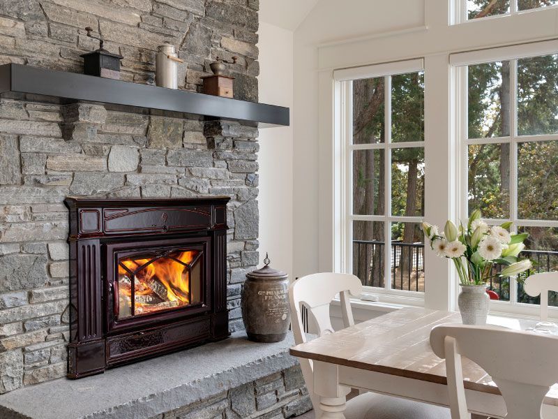 A cozy interior with a stone fireplace, dark cast iron stove, and a dining table. Sunlight streams in from a window.