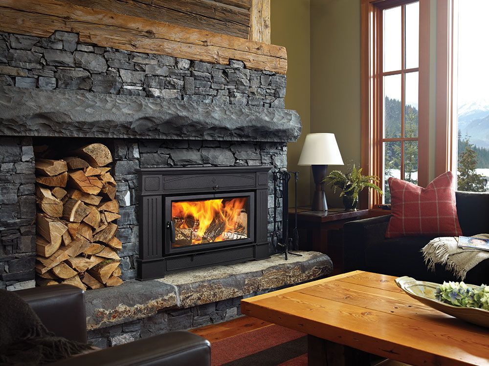A cozy living room with a lit fireplace, stone surround, and stacked firewood. A window offers a scenic view.