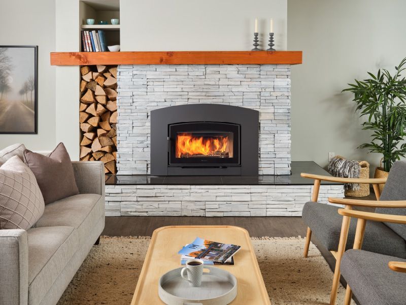 Living room with a lit fireplace built into a stone wall, flanked by a wood stack, a couch, and two chairs.