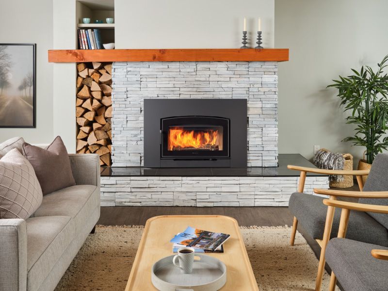 Cozy living room with a fireplace, gray stone surround, wooden mantel, and stacked firewood. A lit fire burns in the insert.