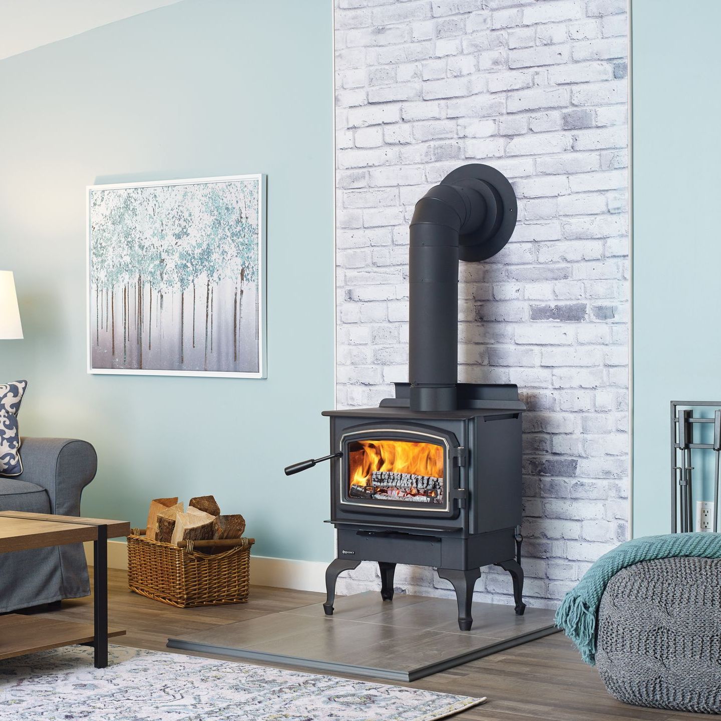A black wood-burning stove with lit fire in a living room, next to a brick-like wall. A painting and wood basket are nearby.