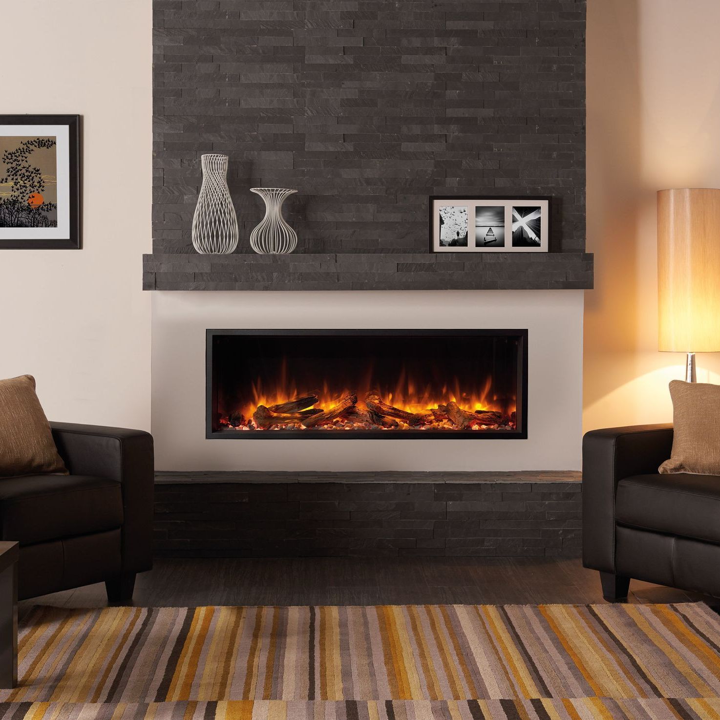 A modern fireplace with a faux flame effect is set into a dark stone wall. Two leather armchairs face it, with a rug below.