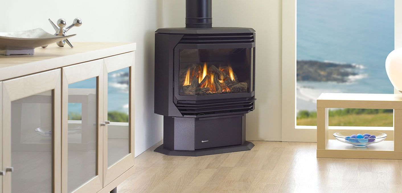A black, freestanding gas fireplace glows in a corner of a room with a view of the ocean through a window.