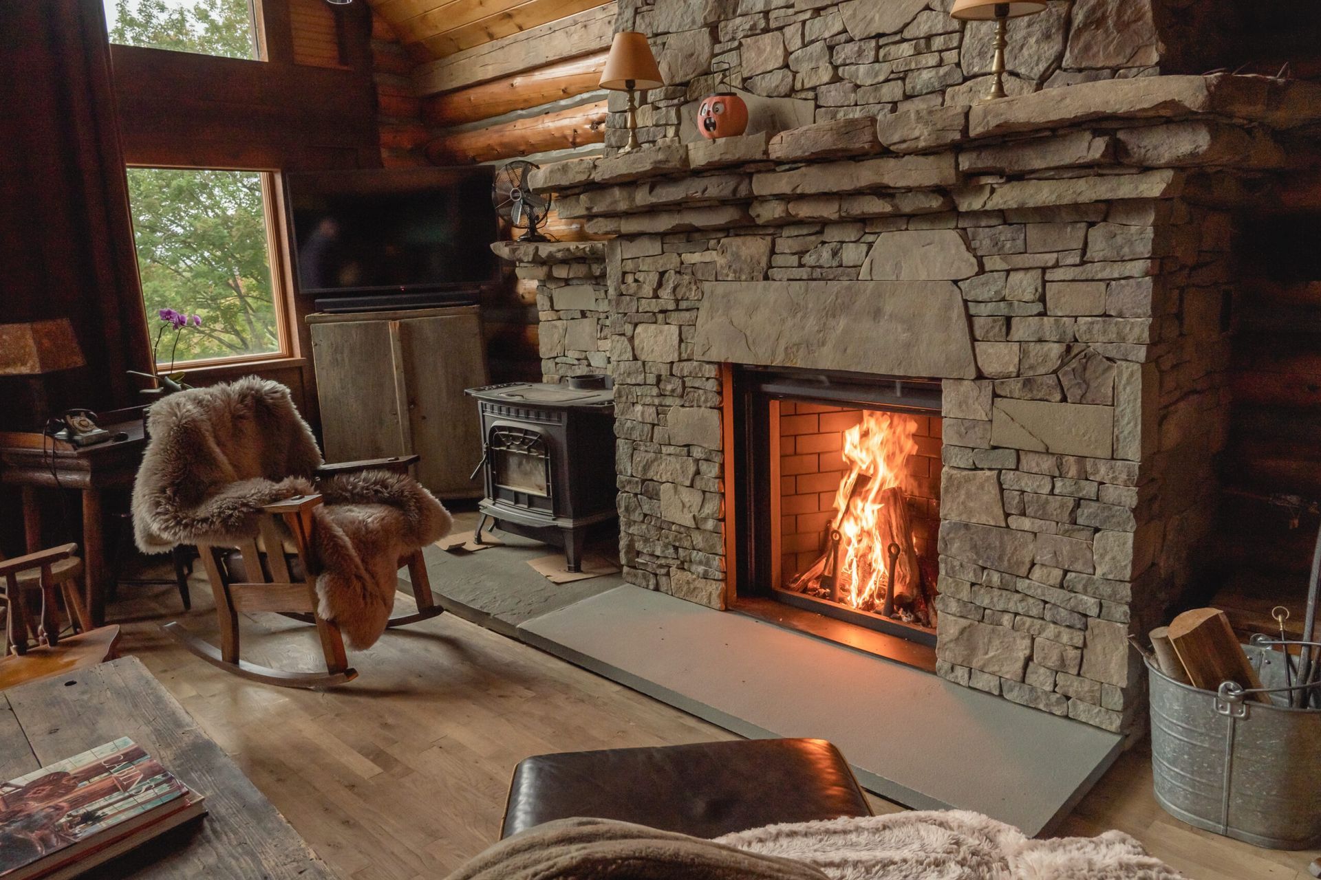 Cozy cabin interior with a lit stone fireplace, rocking chair with fur throw, and logs in a metal bucket.