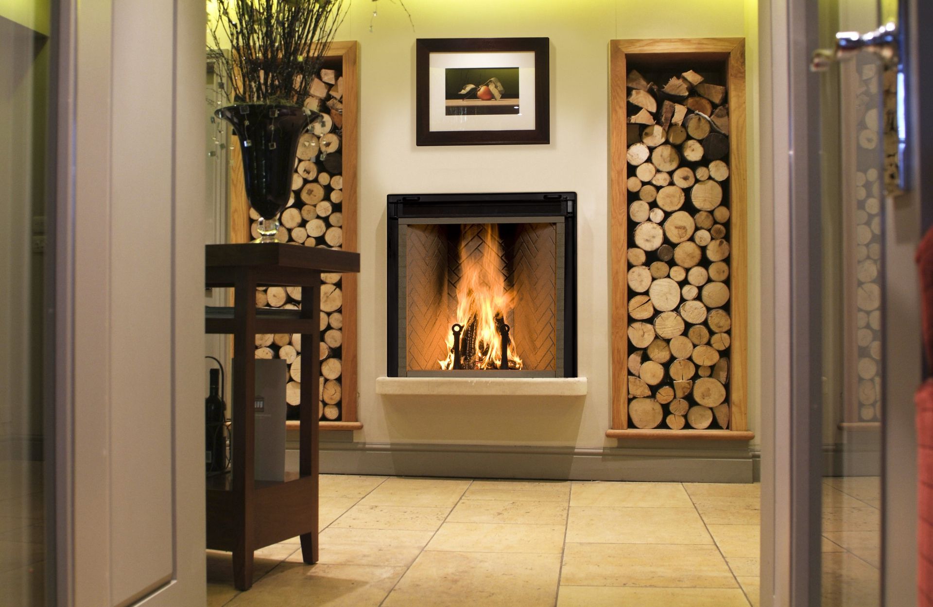 A fireplace in a room with flanking wooden log displays, a small table to the left, and a framed picture above.