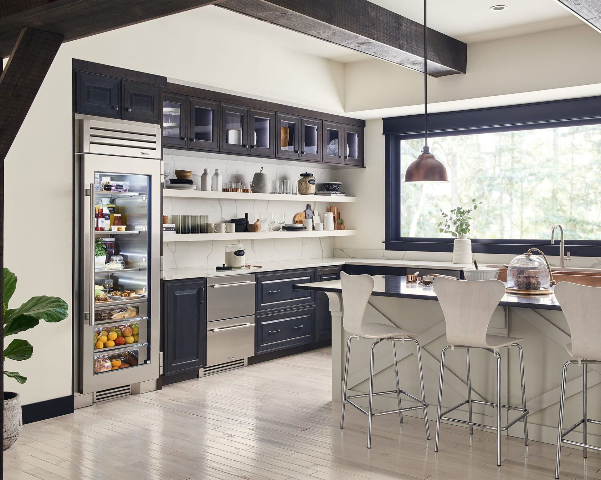 Modern kitchen with stainless steel appliances, dark blue cabinets, white countertops, and bar stools. Large window with a view, light-colored floors.