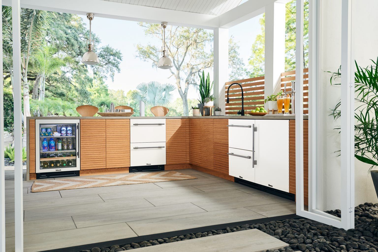Outdoor kitchen with wood cabinets, white appliances, and a beverage cooler under a covered patio. Lush greenery is visible in the background.
