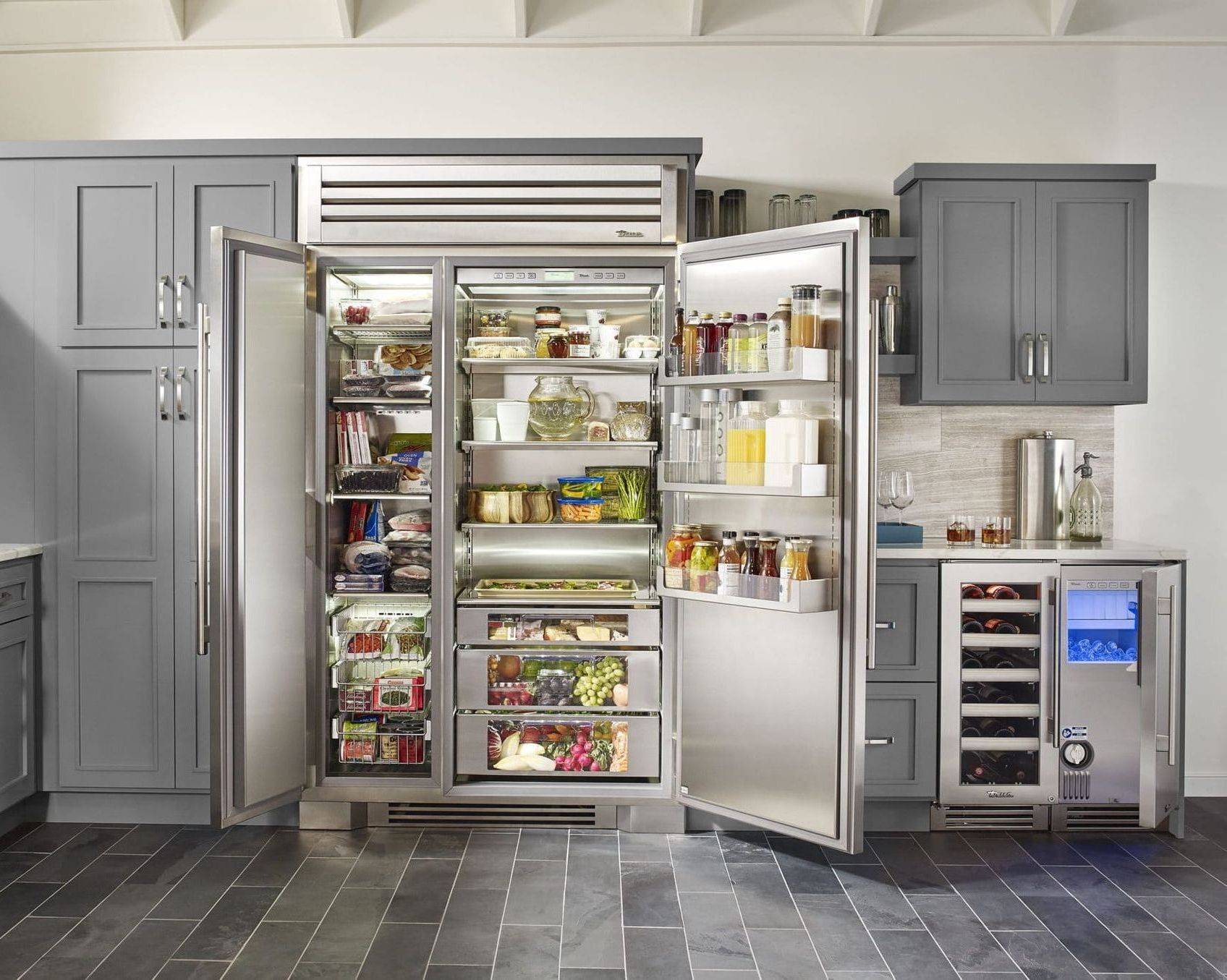 A modern kitchen with an open stainless steel refrigerator showcasing food items. Grey cabinets, and wine chiller complete the setting.