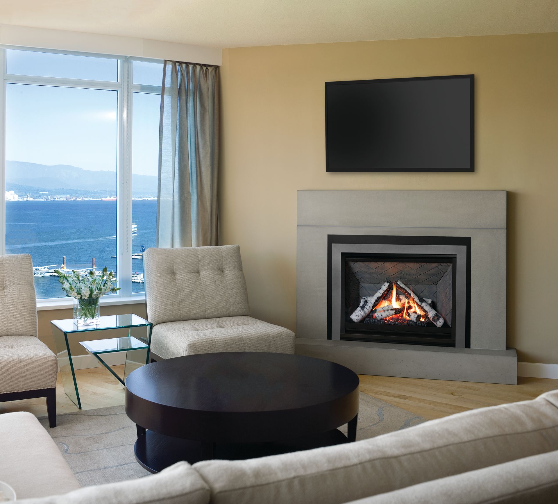 Cozy living room with a fireplace, TV, and a view of the ocean through a large window. There are light-colored chairs and a coffee table.