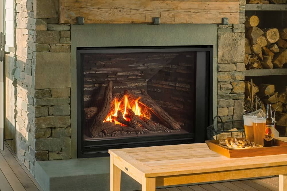 A gas fireplace with burning logs, surrounded by a stone facade, next to a wooden table with drinks and snacks.