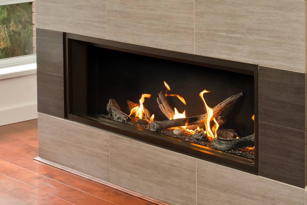A modern fireplace with glowing orange flames and decorative logs, set within a tiled surround of neutral and dark brown tones.