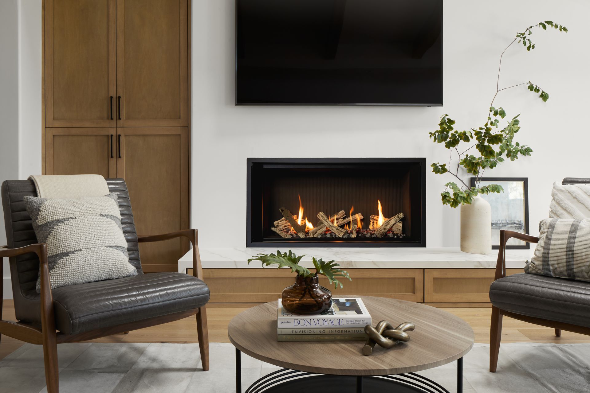 Cozy living room with a fireplace, TV, and two armchairs. A round coffee table sits between them, with decor like plants and books.