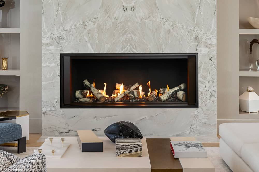 Fireplace in a modern living room with a marble wall, surrounded by shelves and a coffee table. Flames are visible in the fireplace.