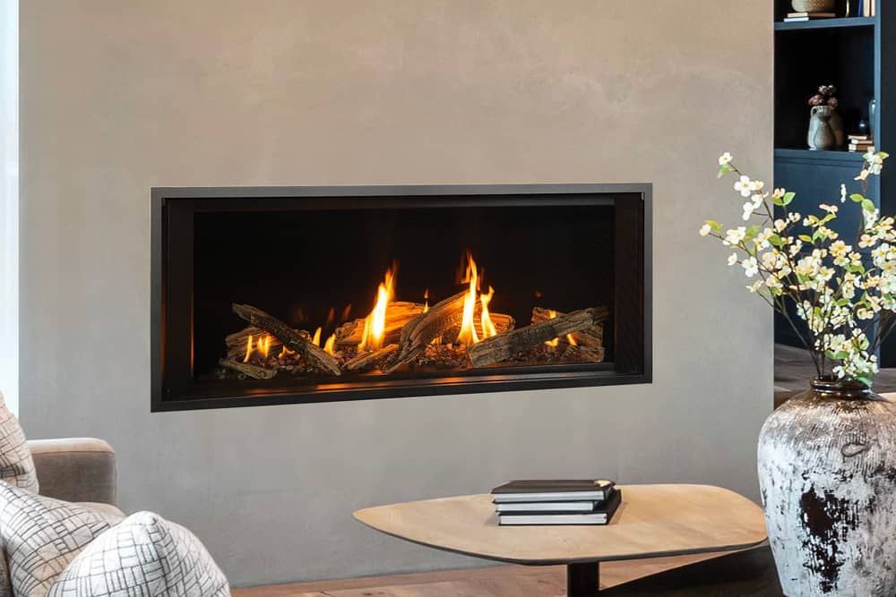 A modern gas fireplace with burning flames and logs built into a gray wall.  A coffee table and vase of flowers are in the foreground.