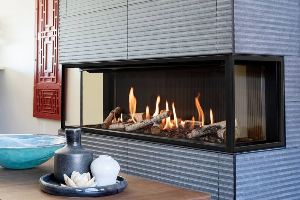 A modern, corner fireplace with a black frame, gas flames, and decorative logs, set against gray tiled walls.  A table with decor is in the foreground.