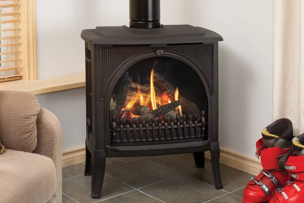 A black gas fireplace with burning flames, logs, and a metal grate, set inside a room with a chair and window.