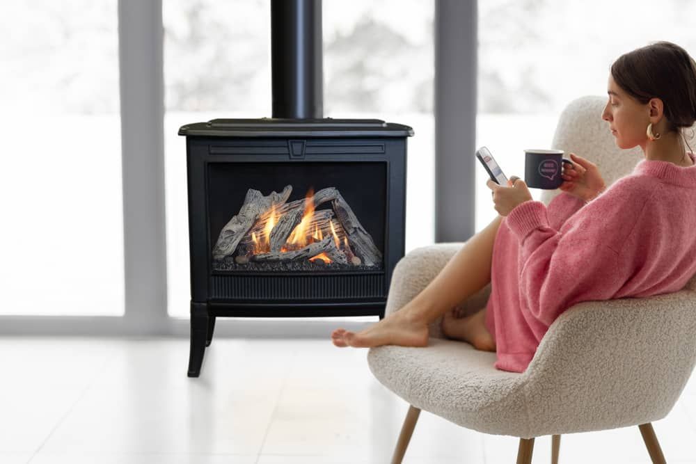 Woman in pink robe, holding mug and phone, sits in a chair by a lit black fireplace in a bright room with a snowy view.