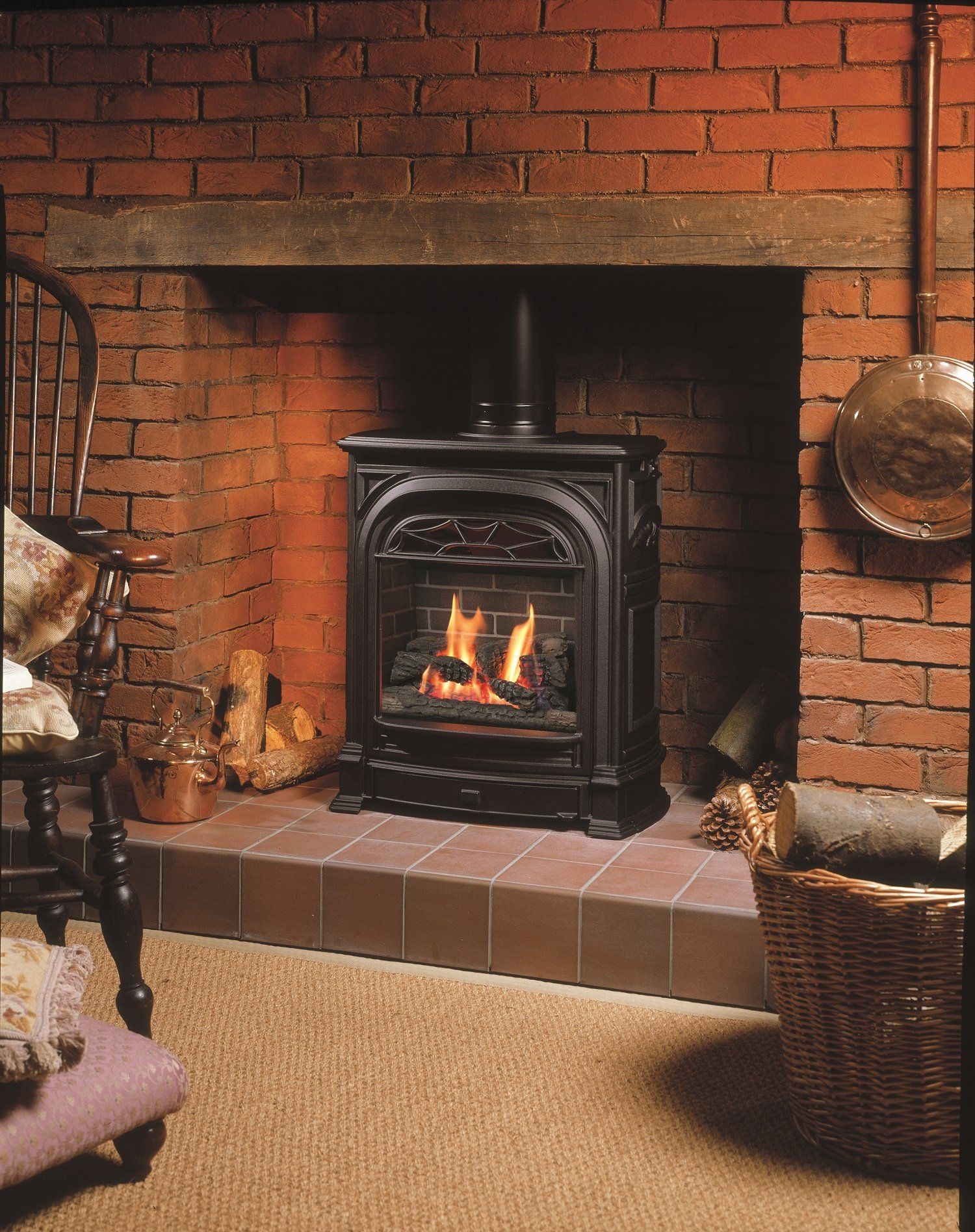 A black, ornate gas fireplace burning in a brick fireplace. A wicker basket with logs sits beside it.