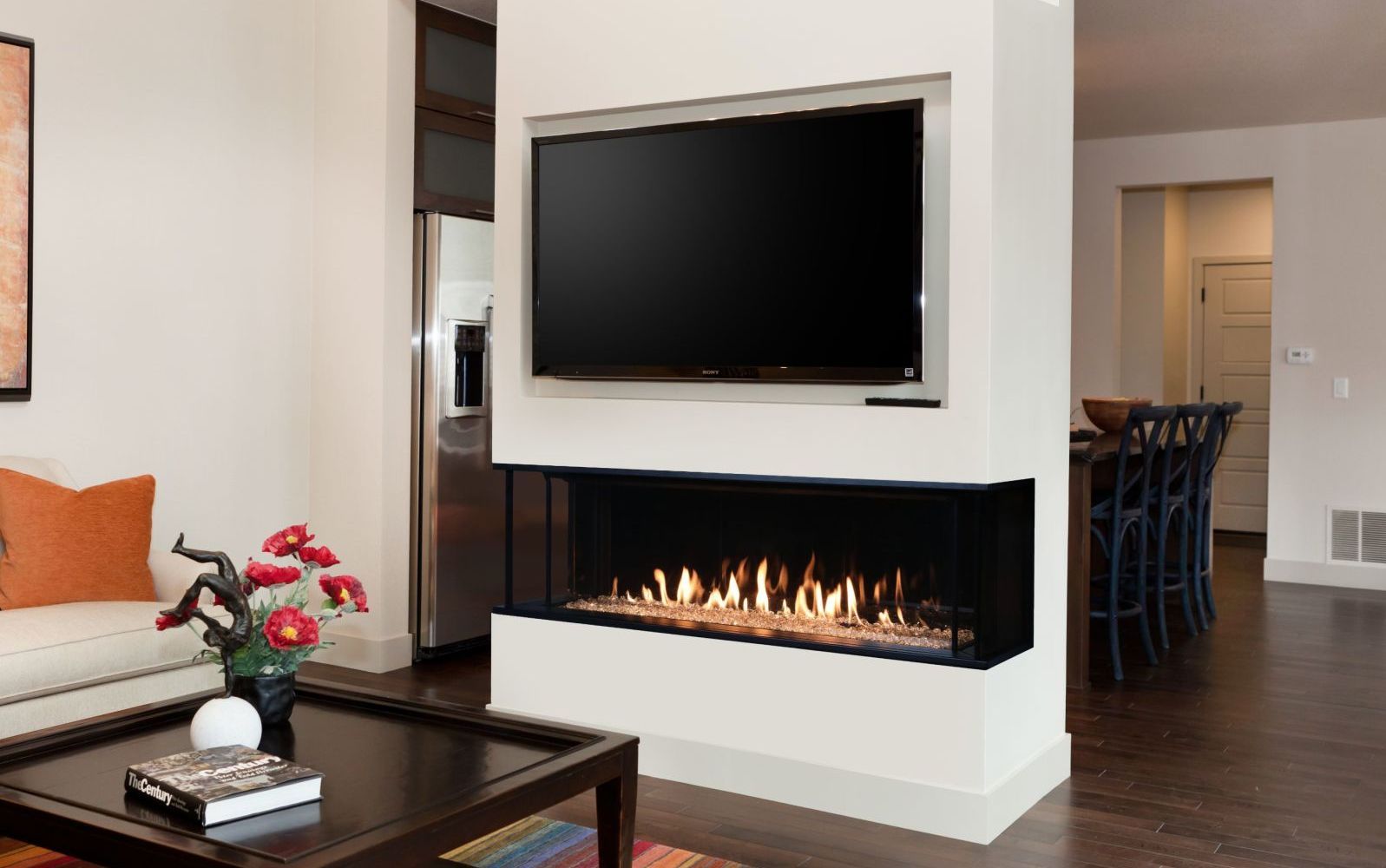 Modern living room with a corner fireplace and mounted TV; white walls, dark wood floors, and a sofa with orange pillows are visible.