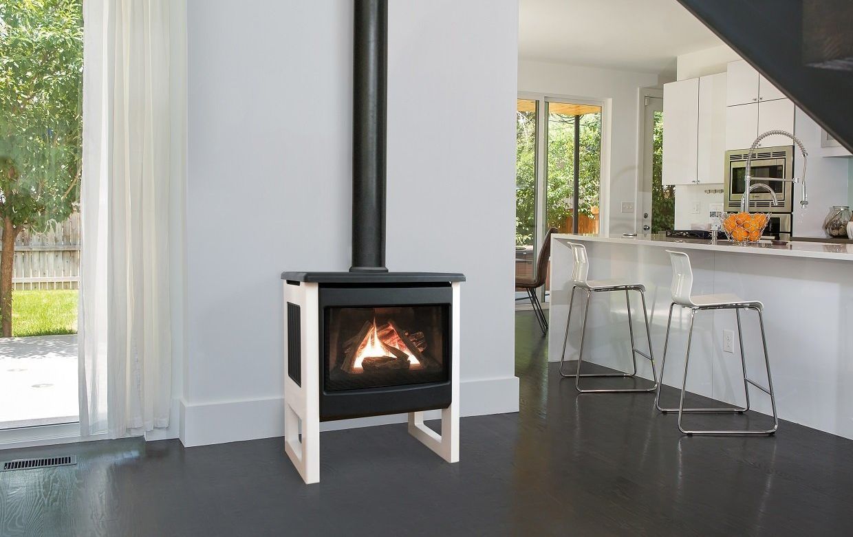 A white and black freestanding gas fireplace with a black chimney pipe stands inside a modern home. The hearth features burning logs, and the kitchen is in the background.