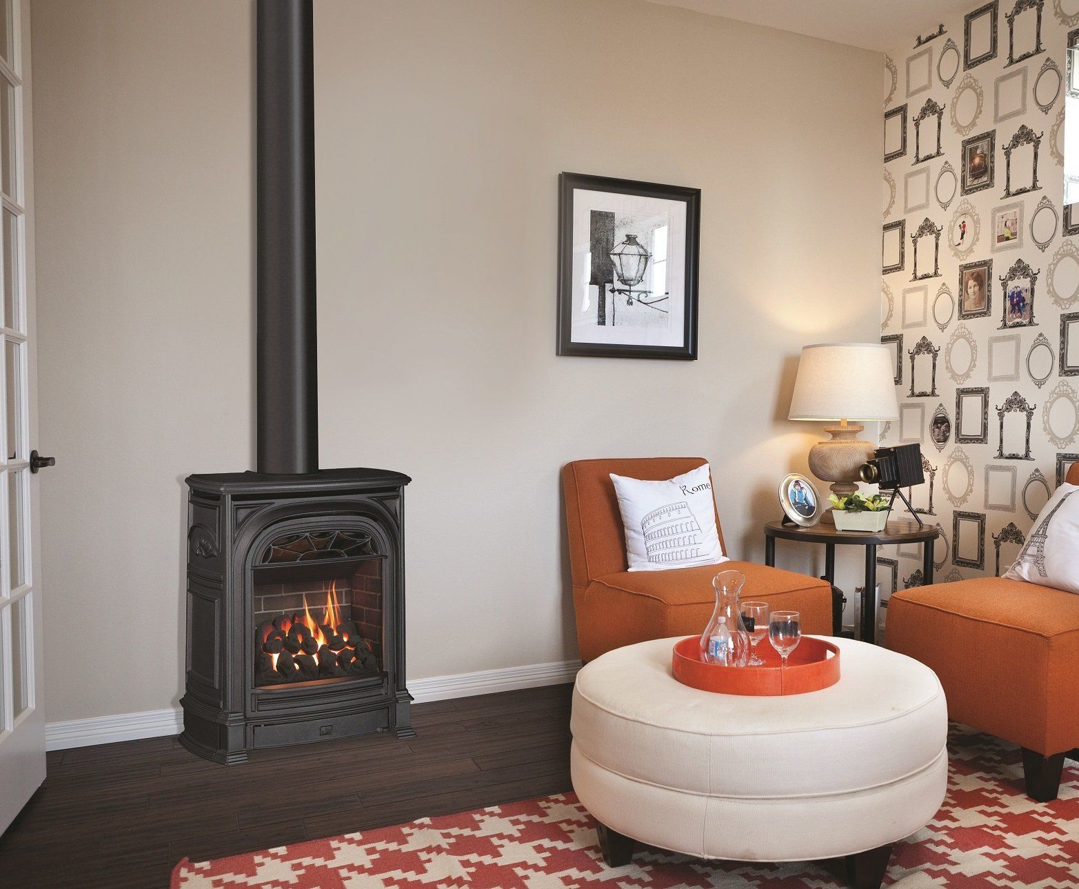 A cozy living room with a fireplace, orange chairs, and a patterned ottoman. The walls are tan and white.