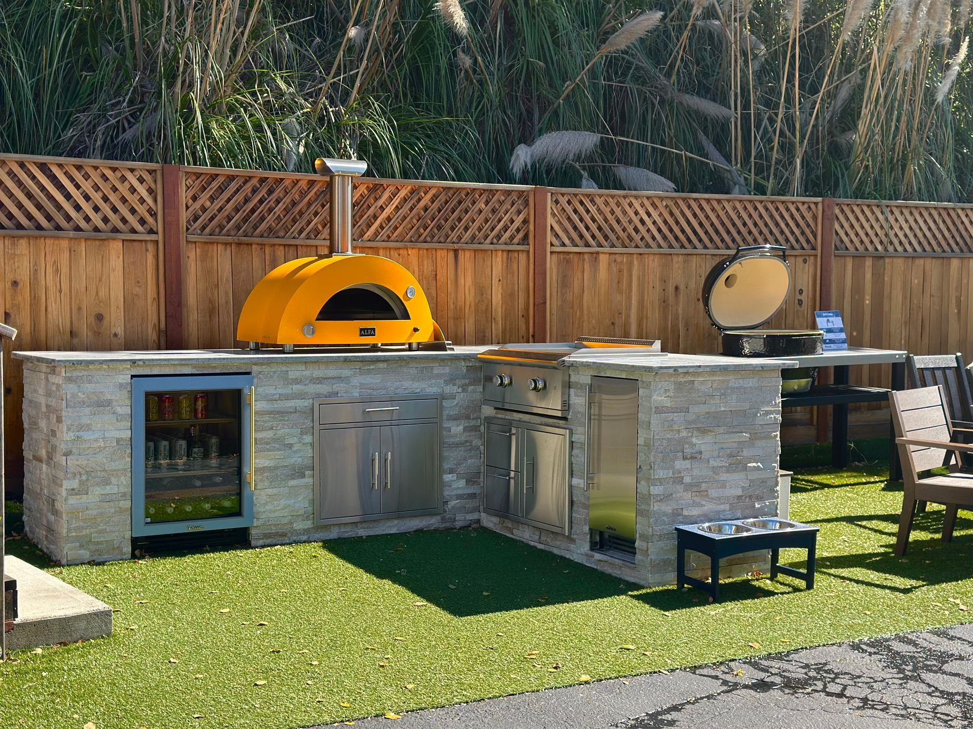 Outdoor kitchen with pizza oven, grill, and fridge on a stone countertop, set against a wooden fence on artificial grass.