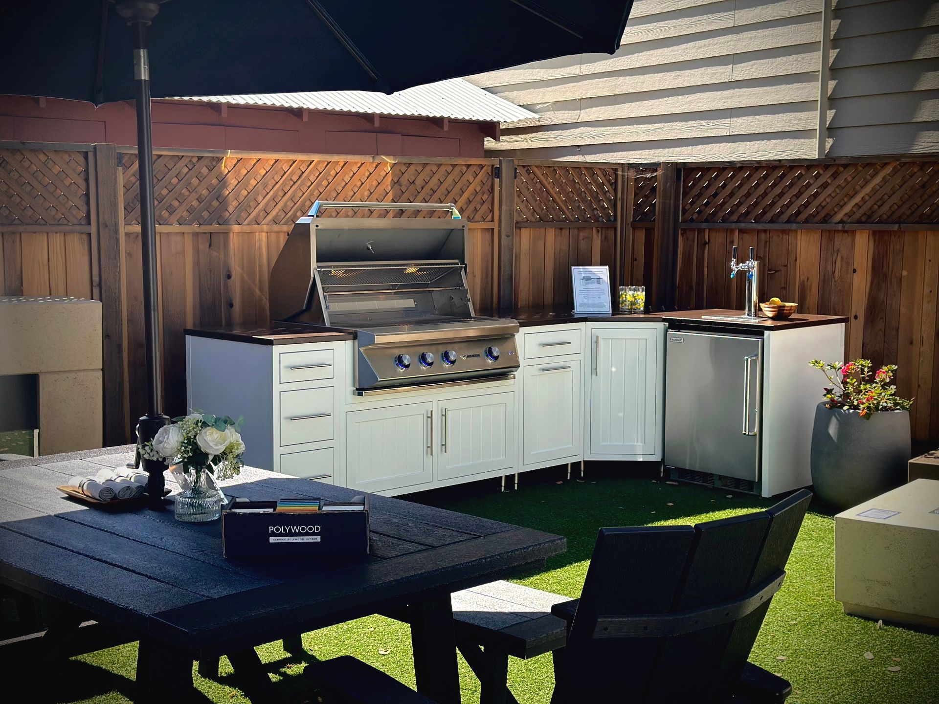 Outdoor kitchen with white cabinets, grill, and refrigerator on a small patch of green turf. A black table and umbrella complete the backyard setup.