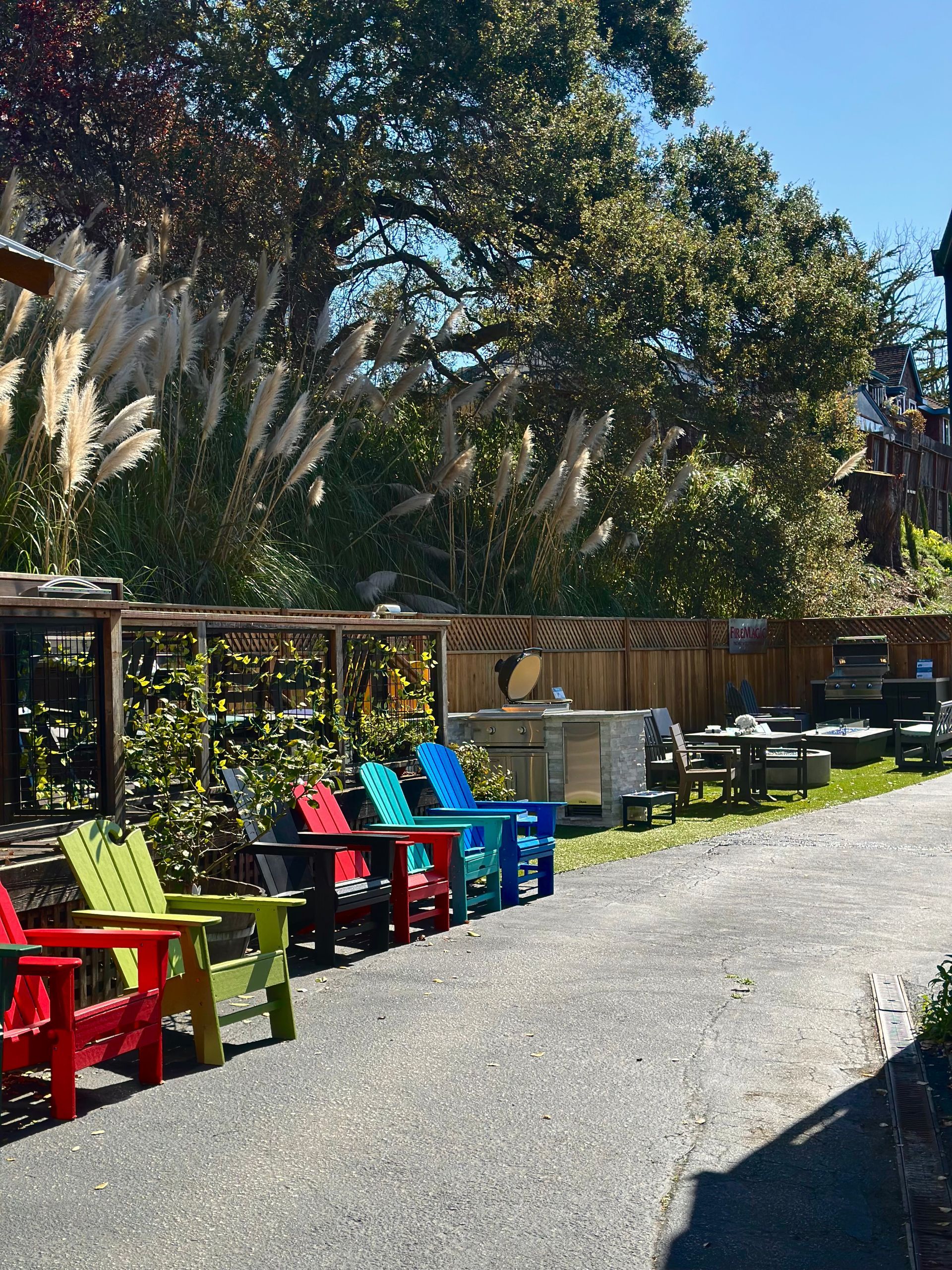 A row of colorful Adirondack chairs faces a gravel path. Tall, feathery pampas grass and trees fill the background.