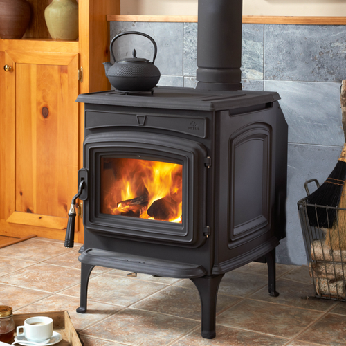 A black wood-burning stove with a lit fire sits on a stone hearth in a log cabin, with a large window overlooking a wooded area.
