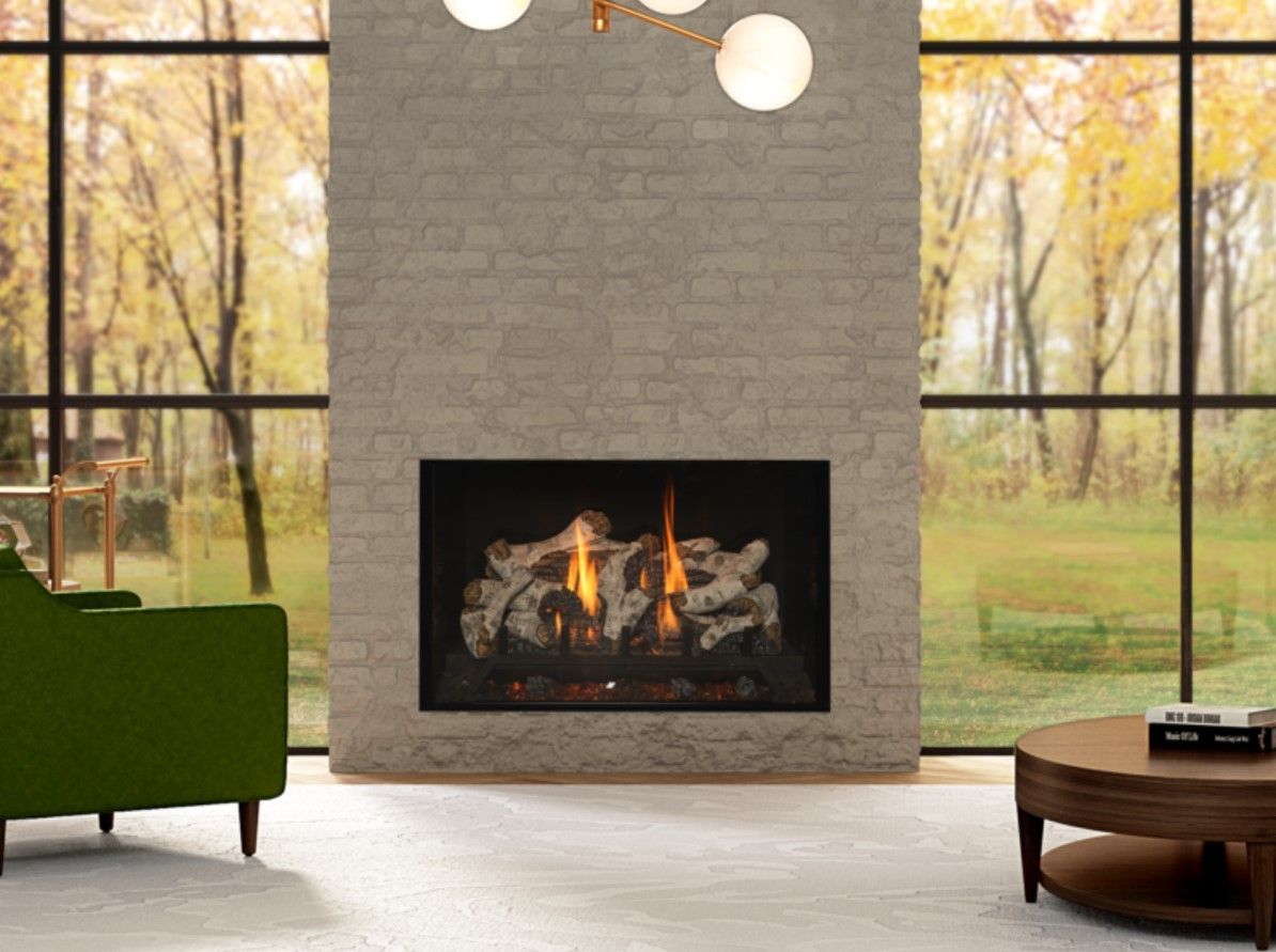 Fireplace in a brick wall with a fire burning, a green chair, and a round table in front of a large window looking onto a fall scene.
