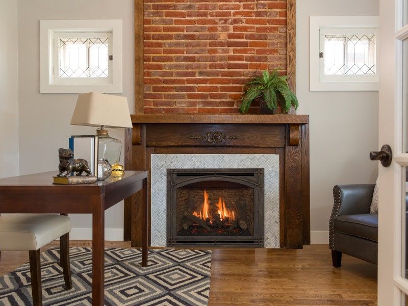 Cozy office with a brick fireplace, wooden desk, and patterned rug. A burning fireplace is visible with a green plant atop.