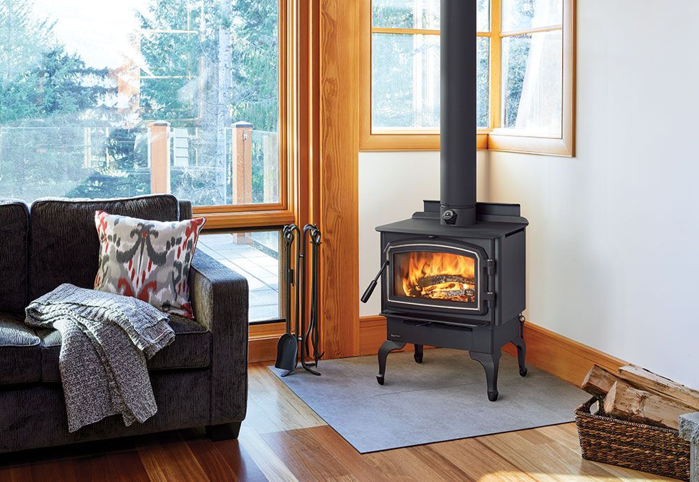 Black wood-burning stove in a corner of a room, logs burning inside. A sofa and window are in the background.