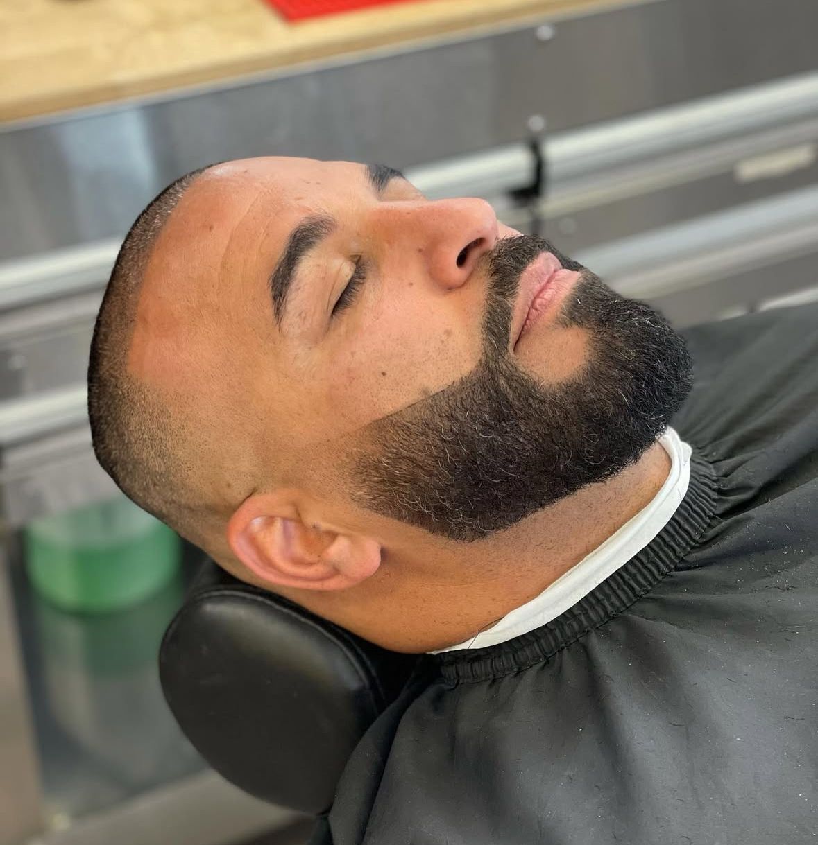 Man reclining in a salon chair with eyes closed, wearing a black cape, sporting a sharp beard fade.