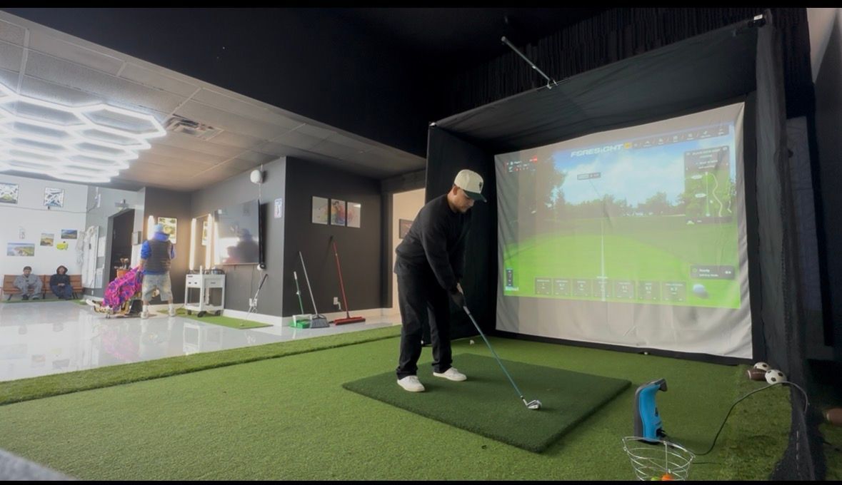 Person golfing indoors in a simulator bay, hitting a ball toward a projected course screen.