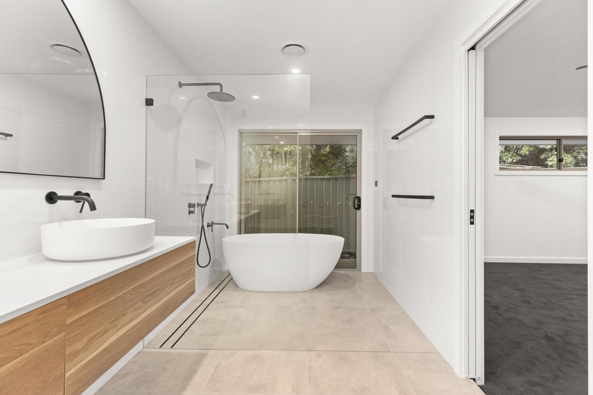 Modern white bathroom with a tub, shower, and vanity. Wooden accents, neutral flooring, and a window to the outside.