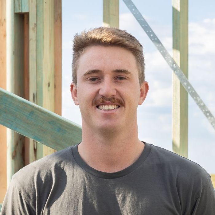 Man with mustache smiling, standing in front of unfinished wooden construction frame. He wears a gray shirt.
