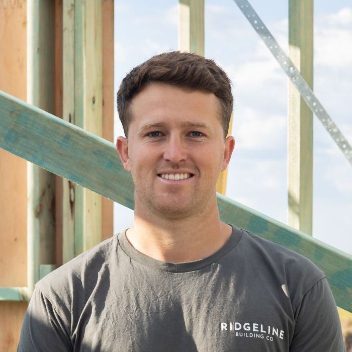 A Brunette Man Wearing a Ridgeline T-shirt is Smiling in Front of a Wooden Structure