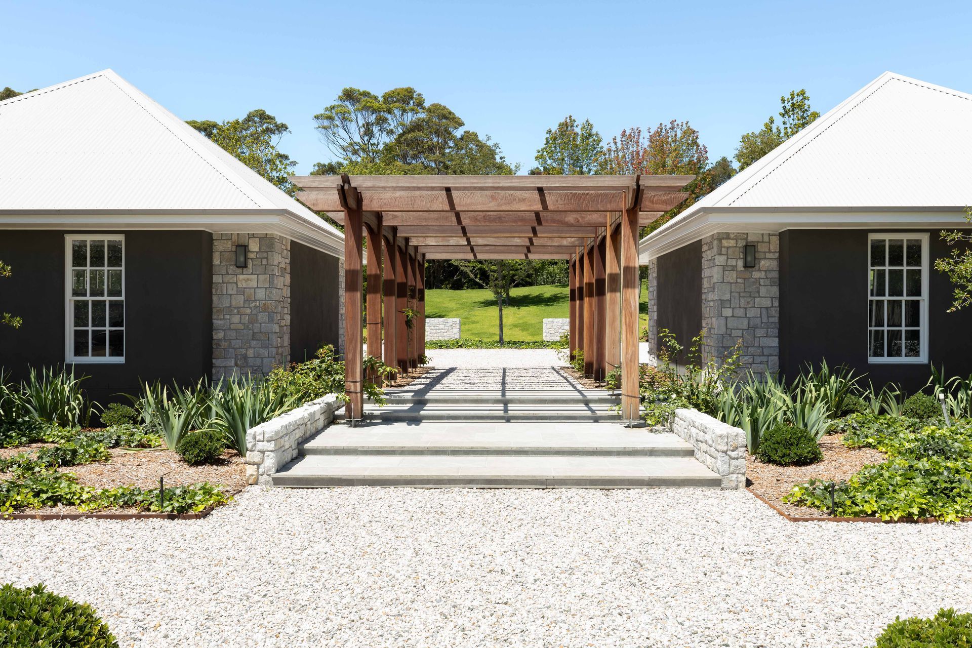 Two dark buildings with white roofs flank a wooden pergola over a stone path.