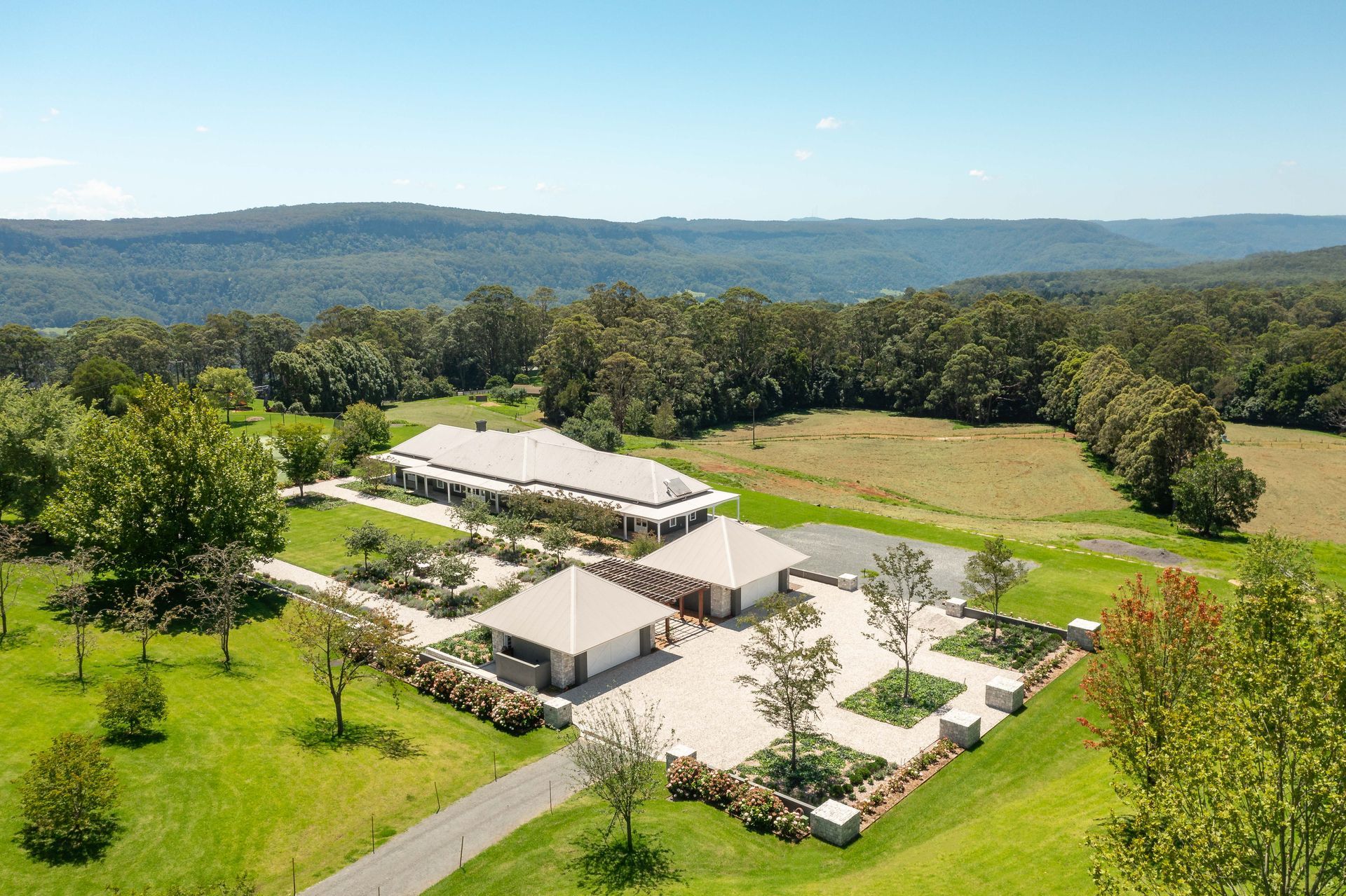 Aerial view of a large house with outbuildings on a grassy hill, surrounded by trees and a distant mountain range.