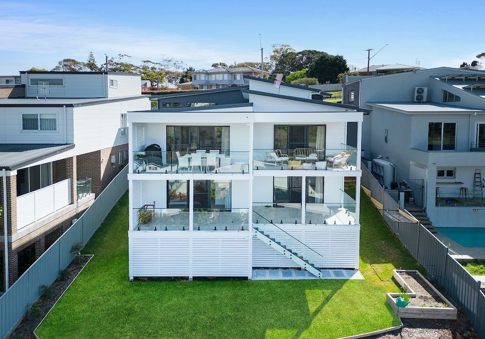 White two-story house with glass balconies, overlooking green lawn and adjacent buildings.