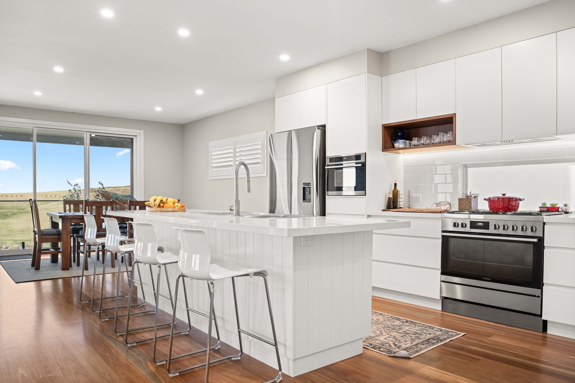 Modern white kitchen with island, stainless steel appliances, and dining area with a view.