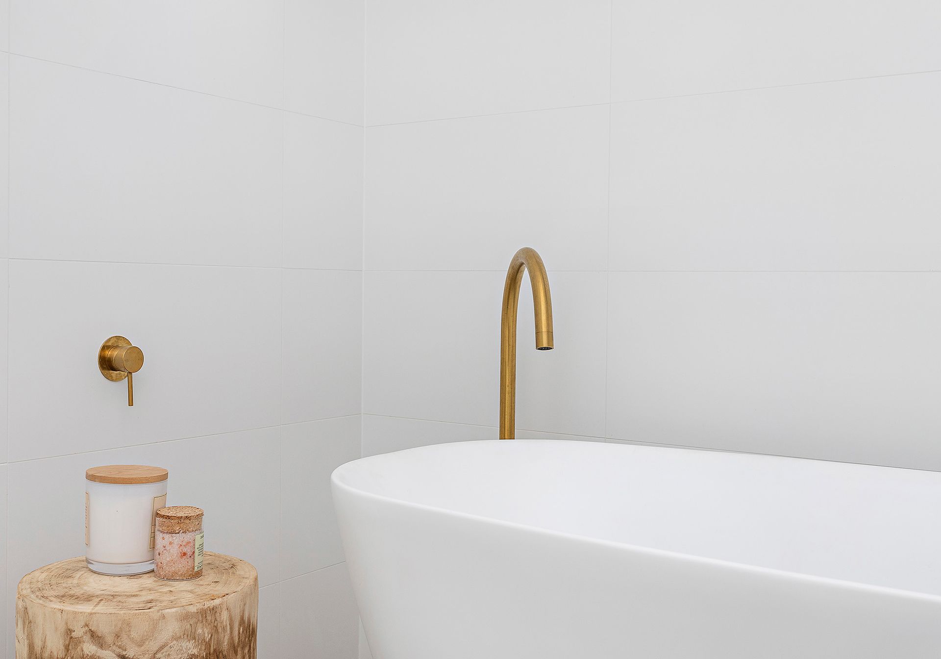 White bathtub with gold faucet and wall fixture. A wooden stool holds candles.