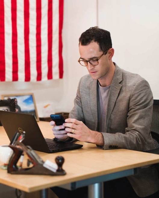 Man in glasses looks at phone, sitting at desk with laptop, American flag in background.