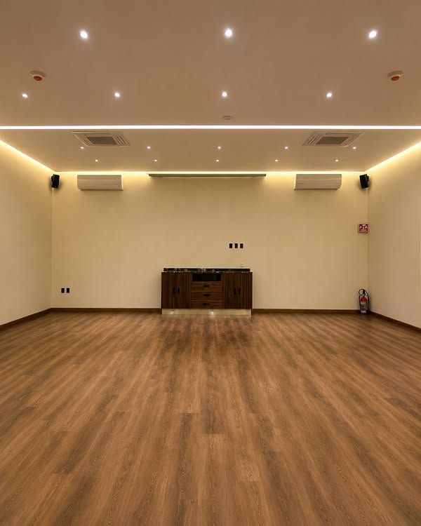 Sala de reuniones vacía con suelo de madera, paredes blancas, luces empotradas y un pequeño armario en la pared del fondo.